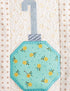 A close-up of a quilted fabric ornament applique featuring a mint green octagonal body with yellow floral print and a light blue stitched handle, set against a cream quilted background with small brown floral motifs.