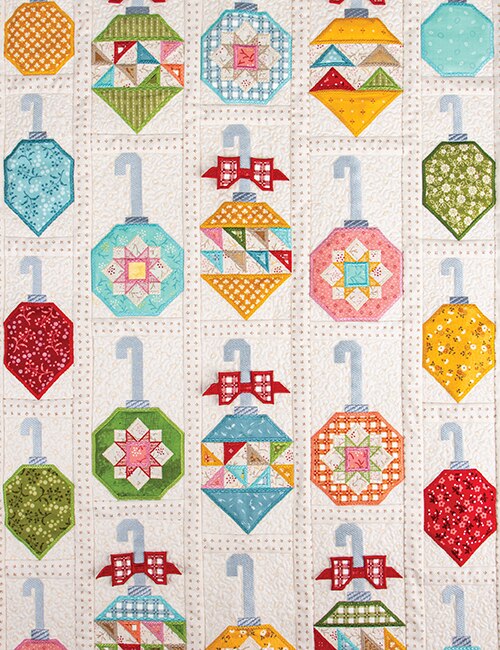A quilted fabric panel featuring a repeating pattern of colorful Christmas ornaments in various geometric shapes and patterns, including stripes, polka dots, and floral prints, hanging from light blue hooks on a cream background.