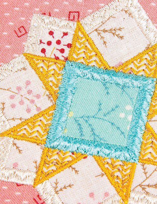 Close-up of a colorful embroidered quilt block featuring a central light blue square surrounded by yellow star-like triangles with white zigzag and floral embroidery on a pink fabric background.