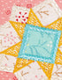 Close-up of a colorful embroidered quilt block featuring a central light blue square surrounded by yellow star-like triangles with white zigzag and floral embroidery on a pink fabric background.