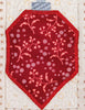 A red fabric ornament-shaped patch with a pink floral and berry pattern, featuring a blue-gray fabric tab at the top, displayed on a cream quilted fabric background.