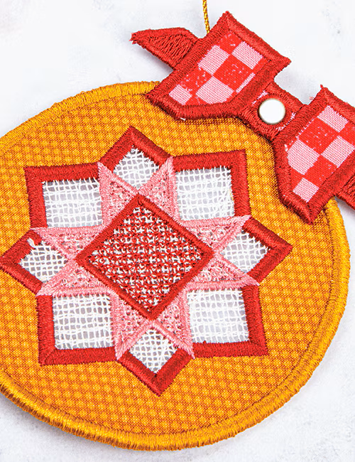 A close-up of a round embroidered textile ornament or decorative patch with a geometric star pattern in red, pink, and white on a yellow-orange stitched background, plus a red-and-white checkered bow detail and a small white snap.