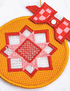 A close-up of a round embroidered textile ornament or decorative patch with a geometric star pattern in red, pink, and white on a yellow-orange stitched background, plus a red-and-white checkered bow detail and a small white snap.