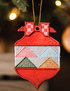 A close-up of a red embroidered Christmas ornament shaped like a house, hanging from a gold string against a softly blurred holiday background with warm lights.