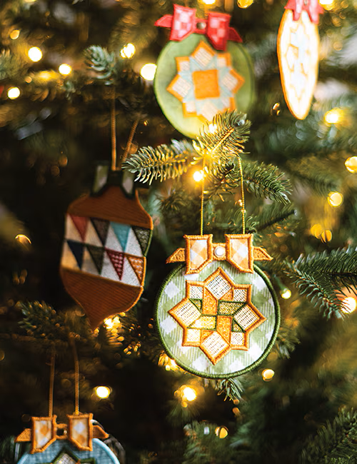 Embroidered quilted fabric Christmas ornaments hanging on a lit Christmas tree. The ornaments feature geometric and starburst patterns in green, orange, white, brown, and red, with warm yellow bokeh lights from the tree lights in the background.
