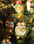 Embroidered quilted fabric Christmas ornaments hanging on a lit Christmas tree. The ornaments feature geometric and starburst patterns in green, orange, white, brown, and red, with warm yellow bokeh lights from the tree lights in the background.