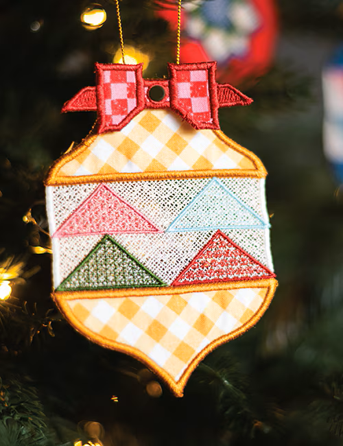 A decorative embroidered Christmas ornament shaped like a holiday tree, hanging on a blurred Christmas tree branch. The ornament features a red checkered bow at the top, a white center band with pink, green, and light blue triangle patterns, and a yellow-and-white checkered lower section with an orange stitched border.
