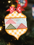 A decorative embroidered Christmas ornament shaped like a holiday tree, hanging on a blurred Christmas tree branch. The ornament features a red checkered bow at the top, a white center band with pink, green, and light blue triangle patterns, and a yellow-and-white checkered lower section with an orange stitched border.