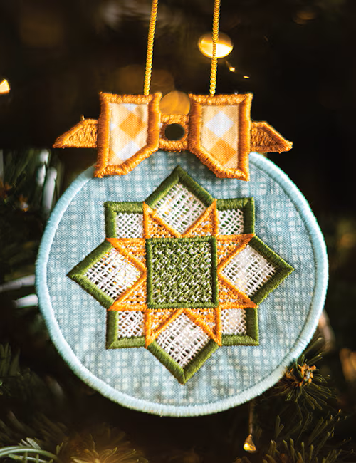A circular embroidered Christmas ornament hanging on a Christmas tree. The ornament features a light blue fabric background with a central starburst design in olive green and orange, topped with two gold bow shapes and hanging from a gold string.