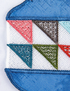 Close-up of a decorative embroidered patch featuring a geometric pattern of triangular flags in pink, brown, teal, green, and red on a white mesh background, bordered by blue embroidered fabric.