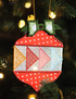 A decorative embroidered Christmas ornament shaped like a strawberry, hanging on a Christmas tree. The ornament features red sections with white polka dots, geometric embroidered patterns in red, yellow, pink, and light blue, and green leaf details at the top.