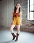 Woman wearing a rust-colored top and yellow shorts with brown boots in an indoor setting.