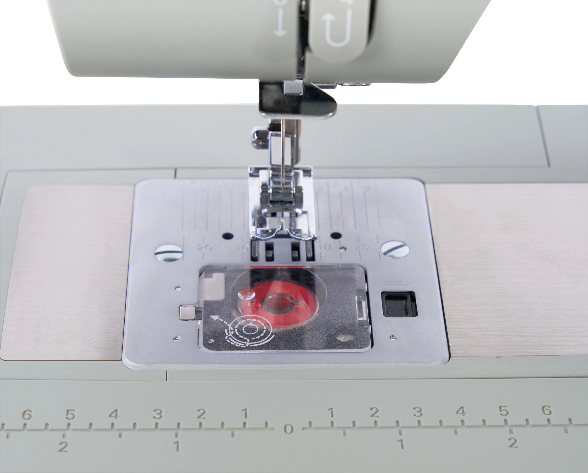 Close-up of a sewing machine needle area and presser foot, showing a transparent cover over a red spool or bobbin component on a light gray machine bed with a ruler scale along the front.