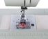 Close-up of a sewing machine needle area and presser foot, showing a transparent cover over a red spool or bobbin component on a light gray machine bed with a ruler scale along the front.