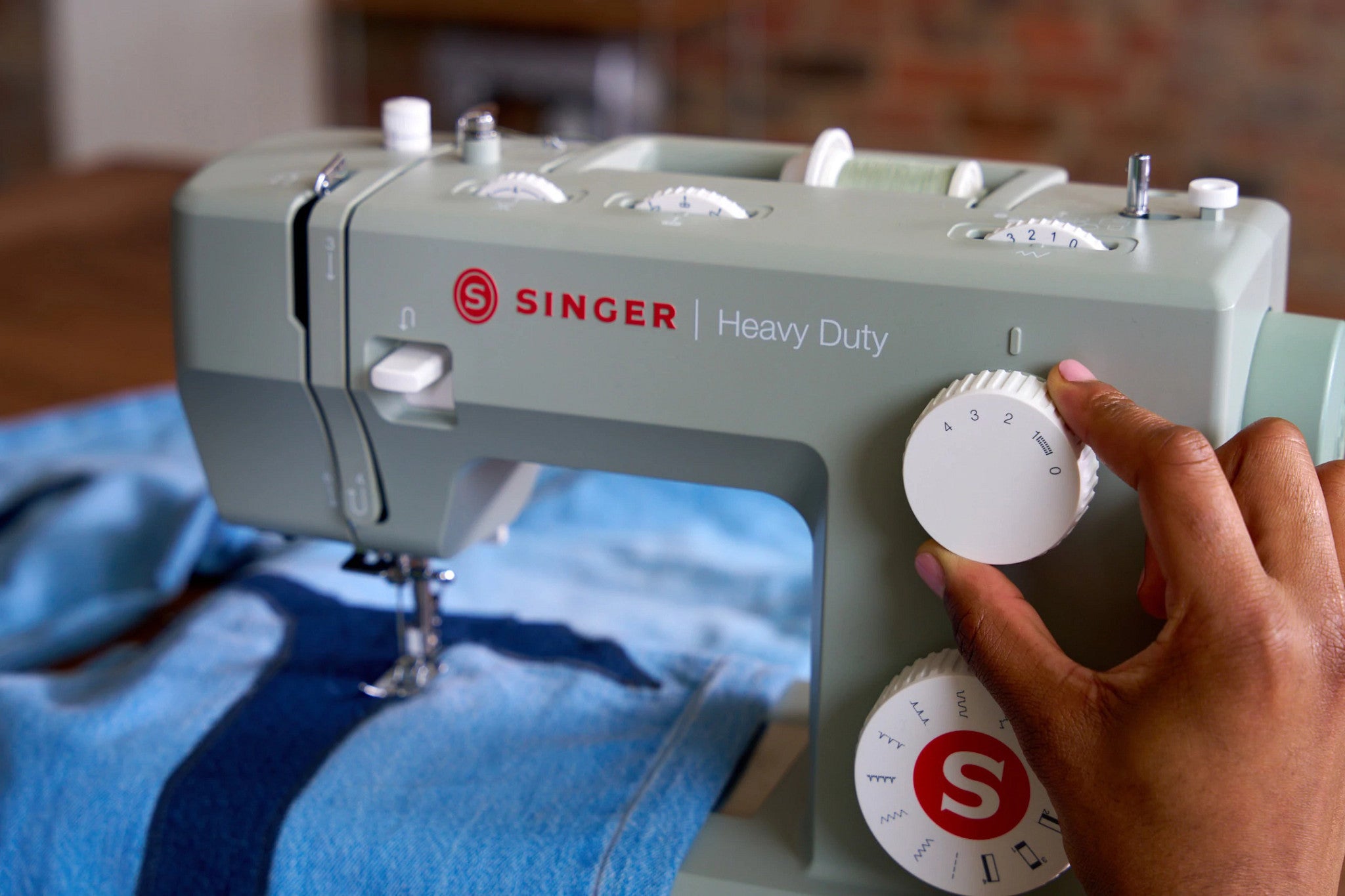 A close-up of a Singer Heavy Duty sewing machine in sage green, with a hand adjusting a white dial. A blue denim garment is visible under the needle, set against a softly blurred indoor background.