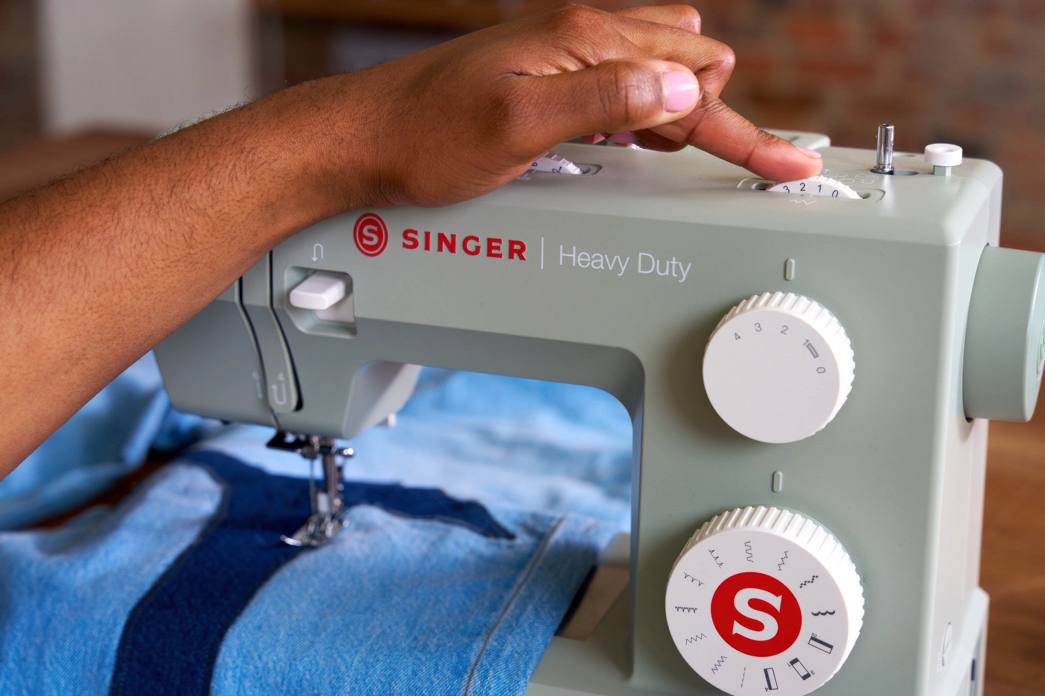 A close-up of a person's hand adjusting the dials of a light sage green Singer Heavy Duty sewing machine while blue denim fabric feeds under the needle.