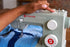 A close-up of a person's hand adjusting the dials of a light sage green Singer Heavy Duty sewing machine while blue denim fabric feeds under the needle.