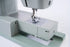 A close-up of a modern sewing machine with a light sage green body, featuring a metallic silver needle plate and presser foot assembly on a clean white background.