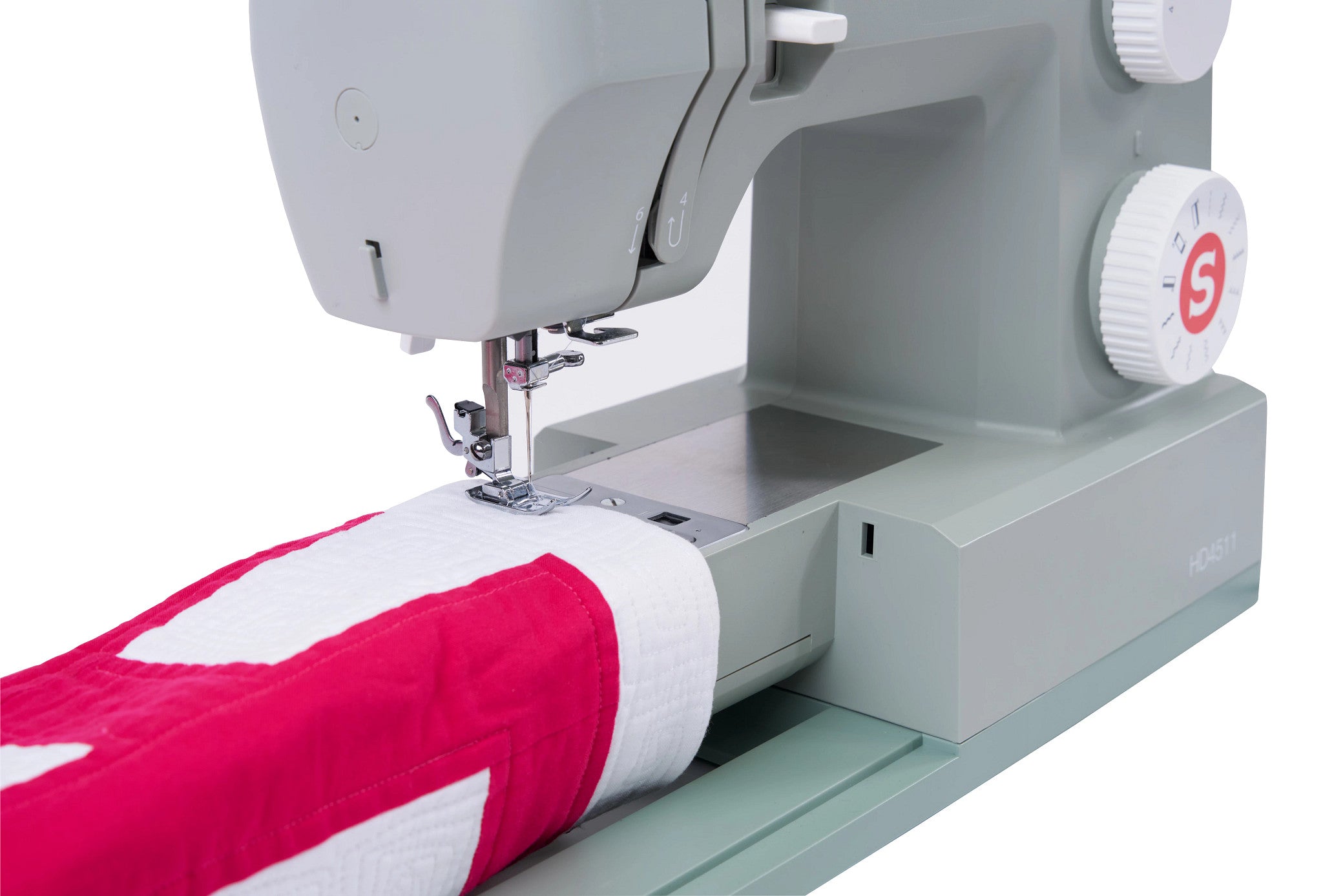 A close-up of a light gray sewing machine with a red and white quilted fabric piece being fed under the needle, shown on a clean white background.