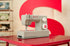 A Singer Mini-Dra sewing machine in light gray sits on a red table, with a large red and cream wall backdrop and other sewing machines blurred in the background.