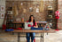 A woman sits at a wooden table with a sewing machine, working in a creative studio with a brick wall background, shelves, and sewing supplies.