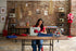 A woman sits at a wooden table in a sewing studio, operating a white sewing machine with blue fabric. The room features a brick wall, shelves with vintage sewing machines and supplies, and a red dress form wearing a patterned dress.