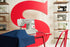 A white Singer sewing machine sits on a red table, sewing blue fabric. A large red letter S is on the wall behind it, with shelves of vintage sewing machines on both sides.