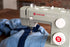 A close-up of a Singer Heavy Duty sewing machine in light gray, shown sewing blue denim fabric. The machine features white dials and a red and white logo on the side, with a blurred indoor background.