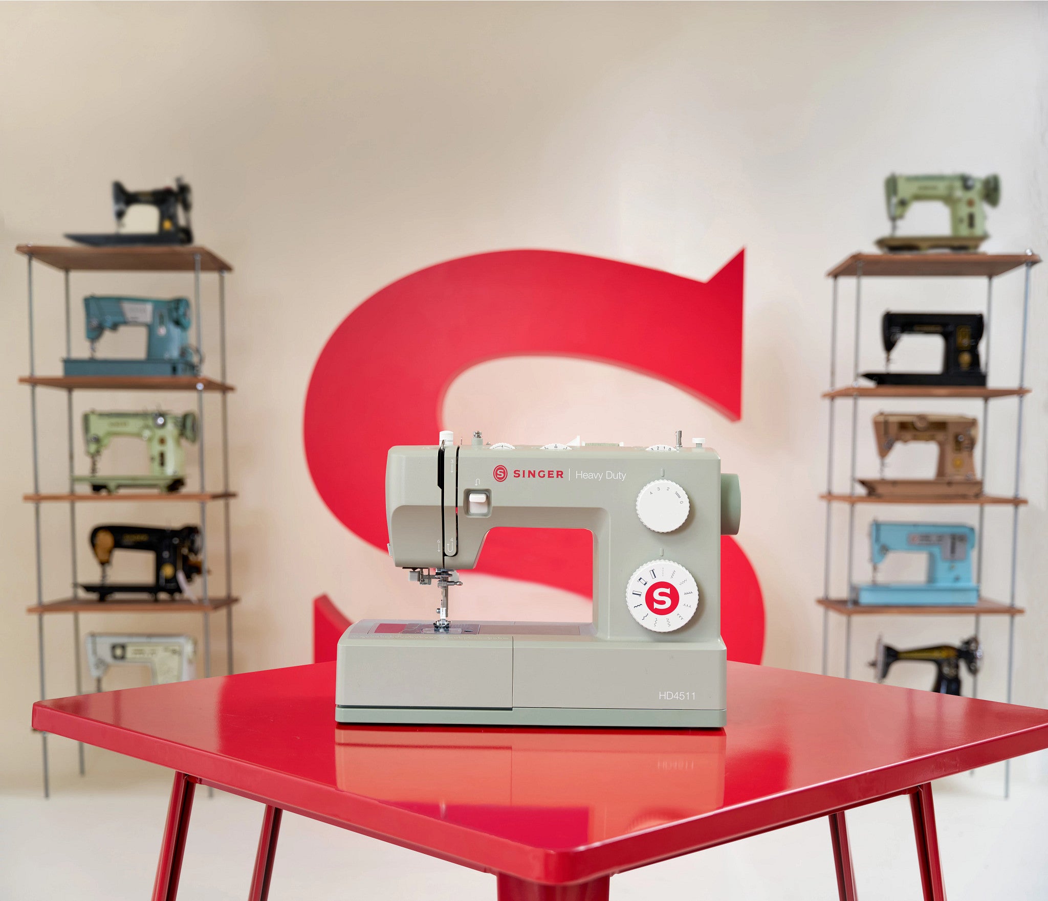 A light gray Singer sewing machine sits on a glossy red table, with a large red letter S on the wall behind it and shelves of vintage sewing machines on both sides.