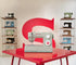 A light gray Singer sewing machine sits on a glossy red table, with a large red letter S on the wall behind it and shelves of vintage sewing machines on both sides.