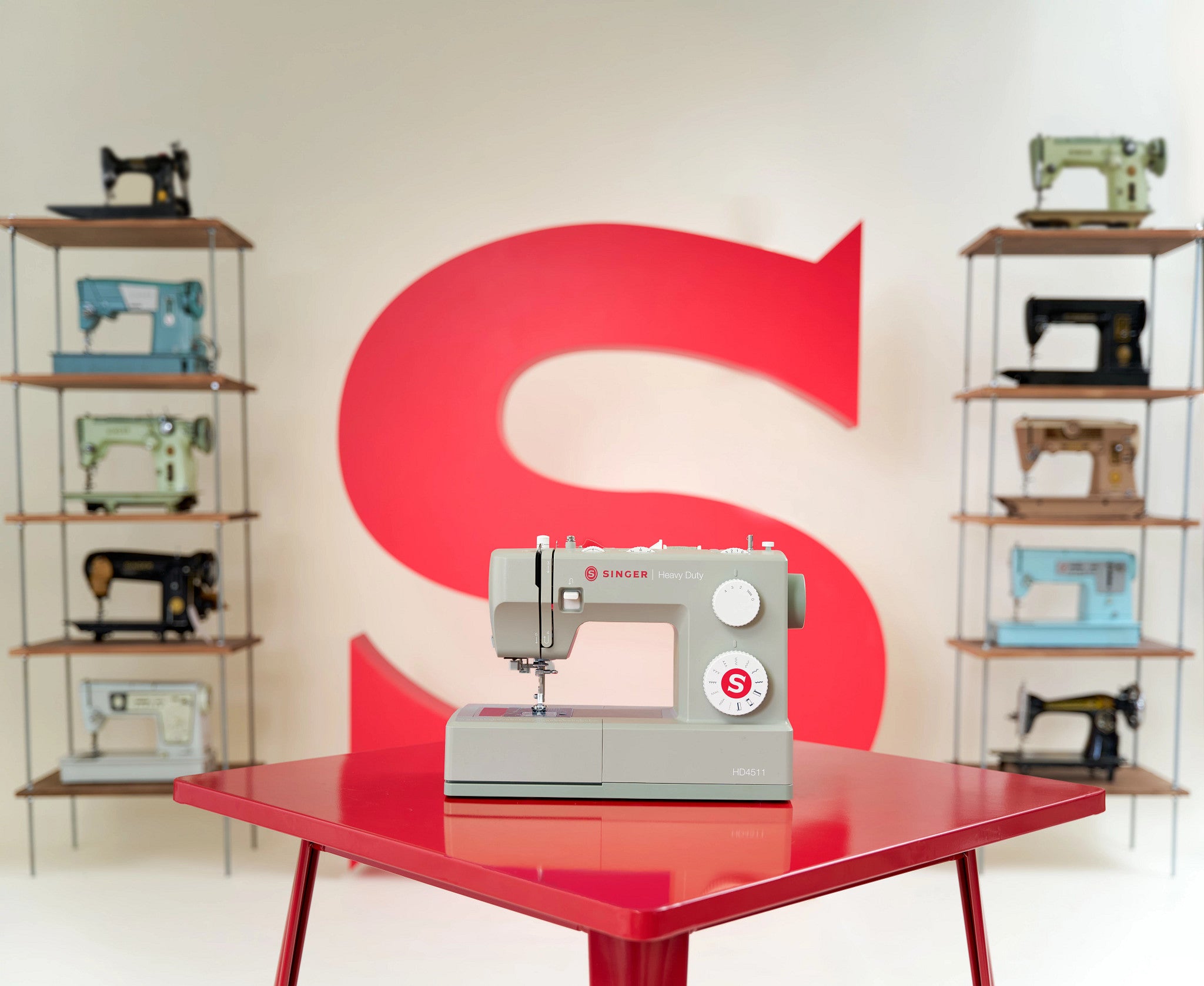 A light gray Singer sewing machine sits on a glossy red table in the foreground, with a large red letter S on the wall behind it and shelves of vintage sewing machines on both sides.