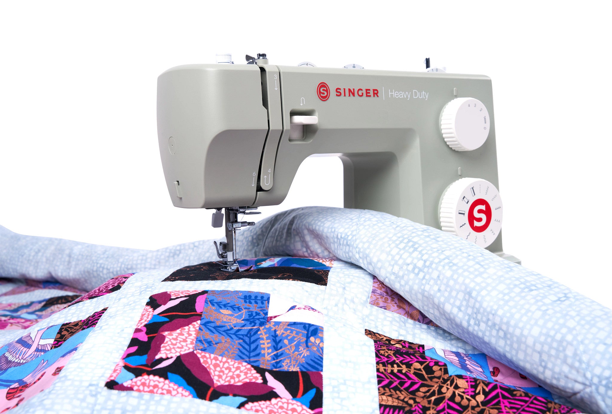 A close-up of a Singer Heavy Duty sewing machine in gray with white dials, stitching a colorful patchwork quilt with light blue fabric, on a white background.