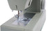 A close-up shot of a sewing machine head and needle area, showing the presser foot, needle, fabric feed dog, and metallic needle plate against a clean white background.