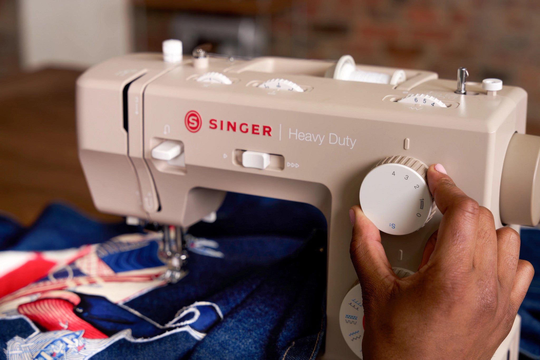 A close-up of a beige Singer Heavy Duty sewing machine with a person's hand adjusting a white dial, with blue denim fabric being sewn underneath.