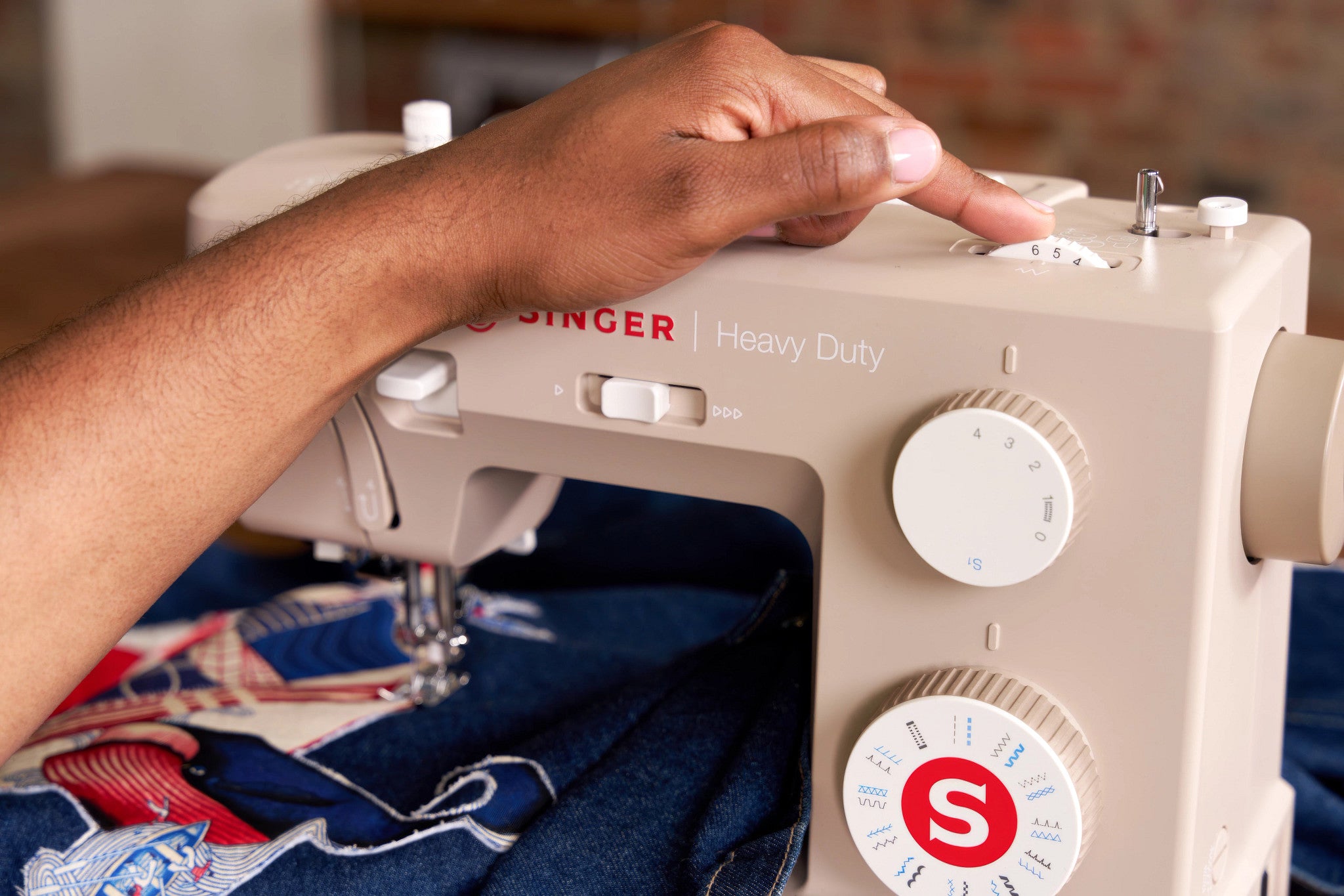 A close-up of a person's hand adjusting the dials of a beige Singer Heavy Duty sewing machine, with blue denim fabric feeding into the needle area.