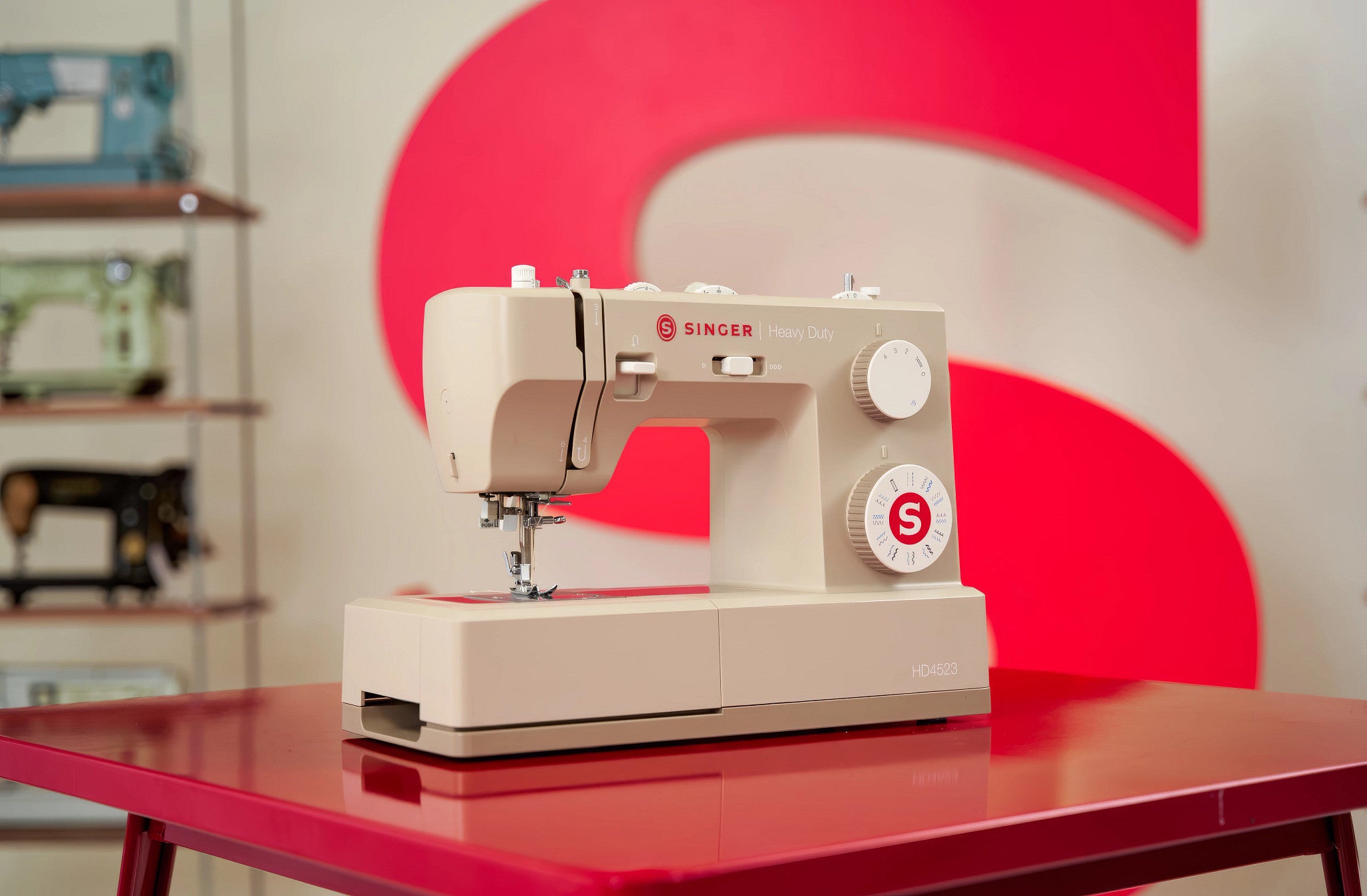 A beige Singer sewing machine sits on a glossy red table, with a large red and white circular backdrop behind it and other sewing machines blurred in the background.