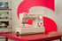 A beige Singer sewing machine sits on a glossy red table, with a large red and white circular backdrop behind it and other sewing machines blurred in the background.
