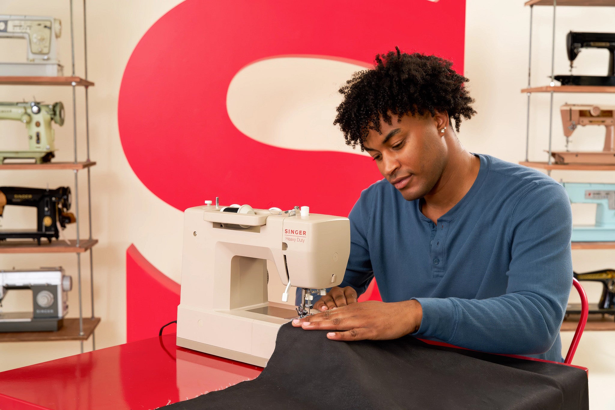 A man in a blue long-sleeve shirt operates a white Singer sewing machine on a red table, with shelves of vintage sewing machines and a large red letter 'S' in the background.