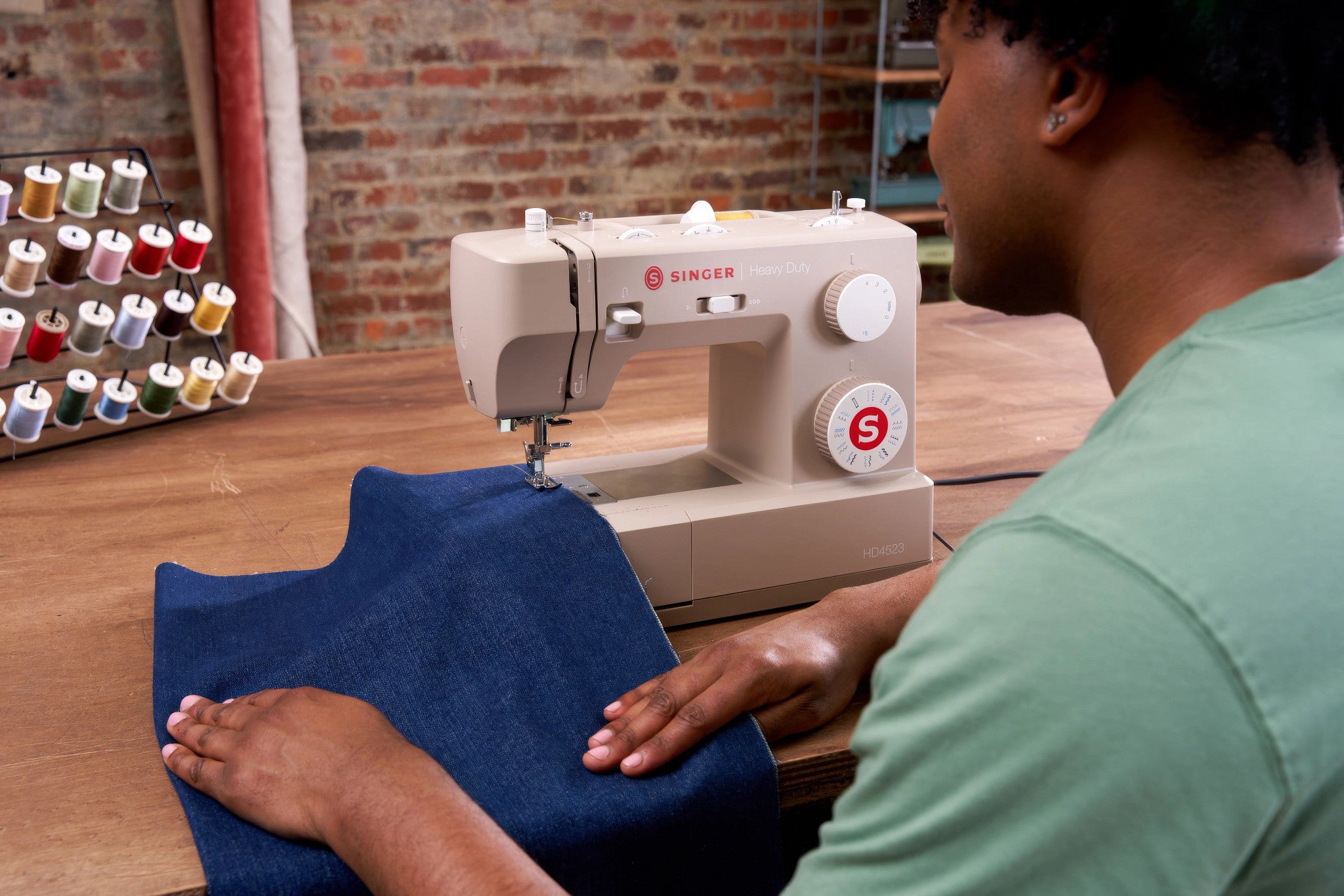 A person in a green shirt guides a navy blue fabric under the needle of a beige Singer sewing machine on a wooden table, with a rack of thread spools and a brick wall in the background.