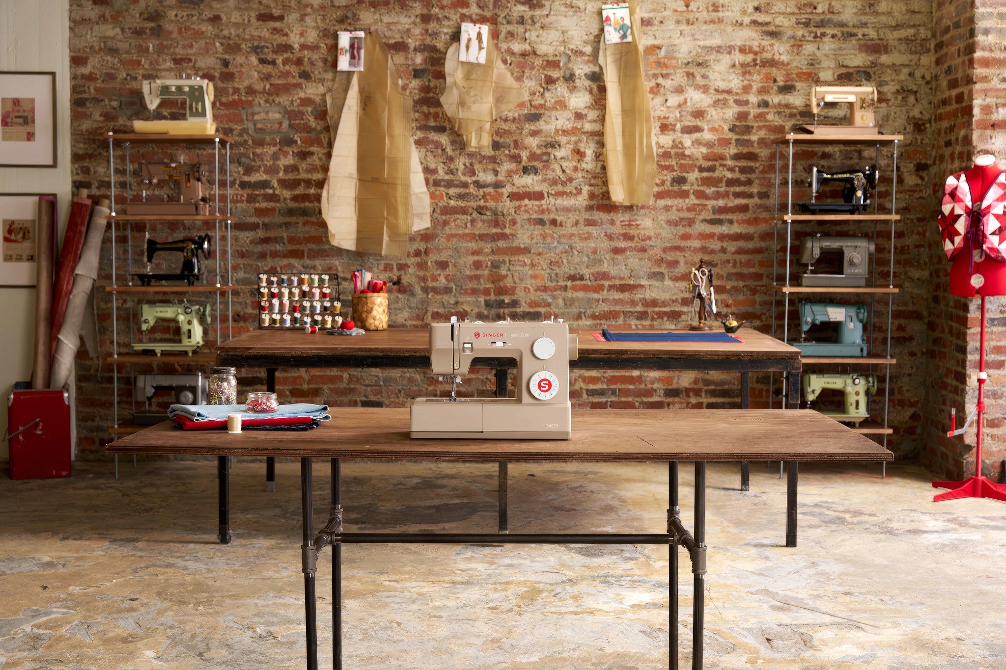 A vintage-style sewing machine sits on a wooden worktable in a rustic sewing room with a brick wall, shelves, and other sewing equipment.