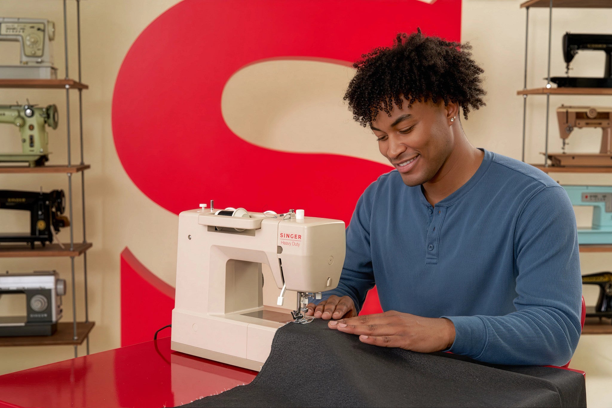 A man in a blue long-sleeve shirt operates a white Singer sewing machine, guiding dark gray fabric under the needle. The background features a large red 'S' logo and shelves displaying vintage sewing machines.