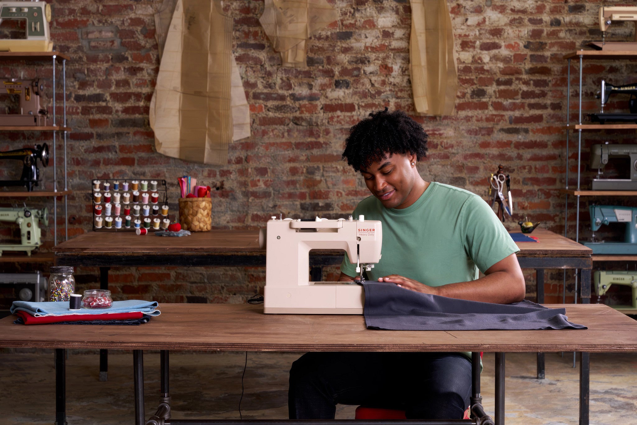 A person sitting at a wooden table in a sewing studio, using a white sewing machine to sew dark fabric. The background features a brick wall, shelves with sewing equipment, and hanging material samples.