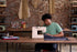 A person sitting at a wooden table in a sewing studio, using a white sewing machine to sew dark fabric. The background features a brick wall, shelves with sewing equipment, and hanging material samples.