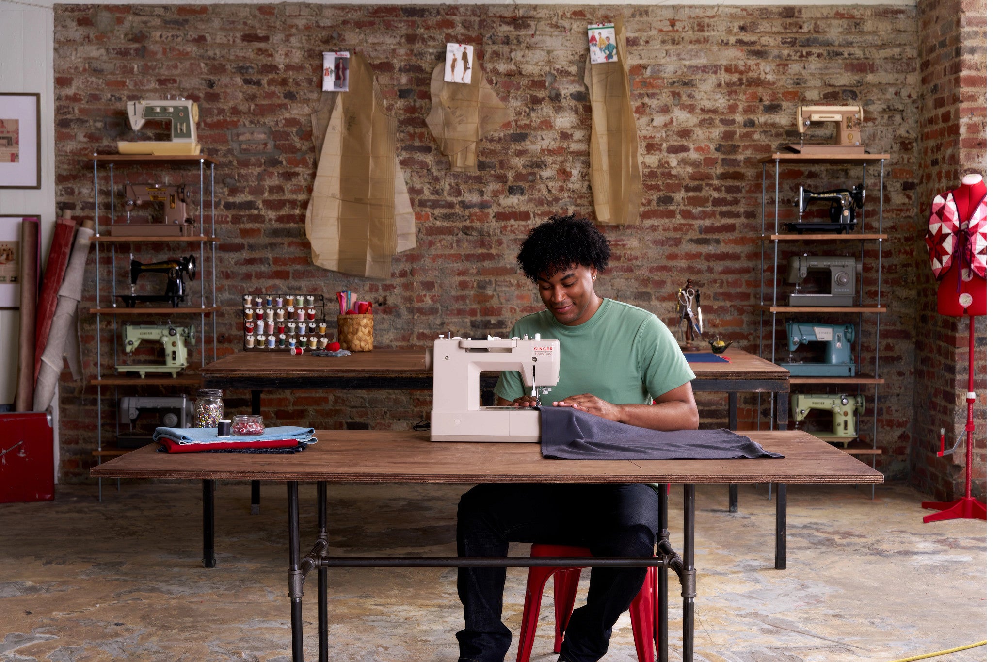 A man in a green t-shirt sits at a wooden table in a sewing studio, operating a white sewing machine with gray fabric. The background features a rustic brick wall with hanging fabric samples, shelves displaying vintage sewing machines, and a red dress form on the right.