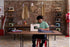 A man in a green t-shirt sits at a wooden table in a sewing studio, operating a white sewing machine with gray fabric. The background features a rustic brick wall with hanging fabric samples, shelves displaying vintage sewing machines, and a red dress form on the right.