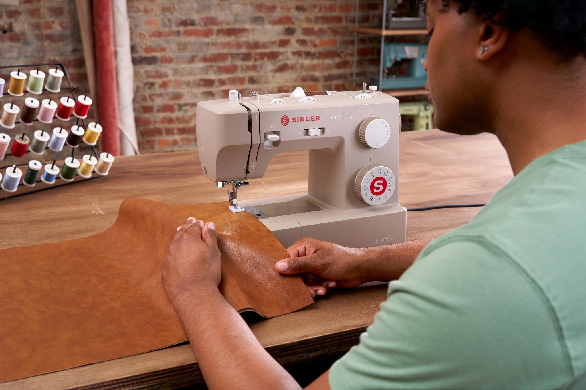 A person using a beige Singer sewing machine to sew brown leather fabric on a wooden table, with a brick wall and spools of thread in the background.