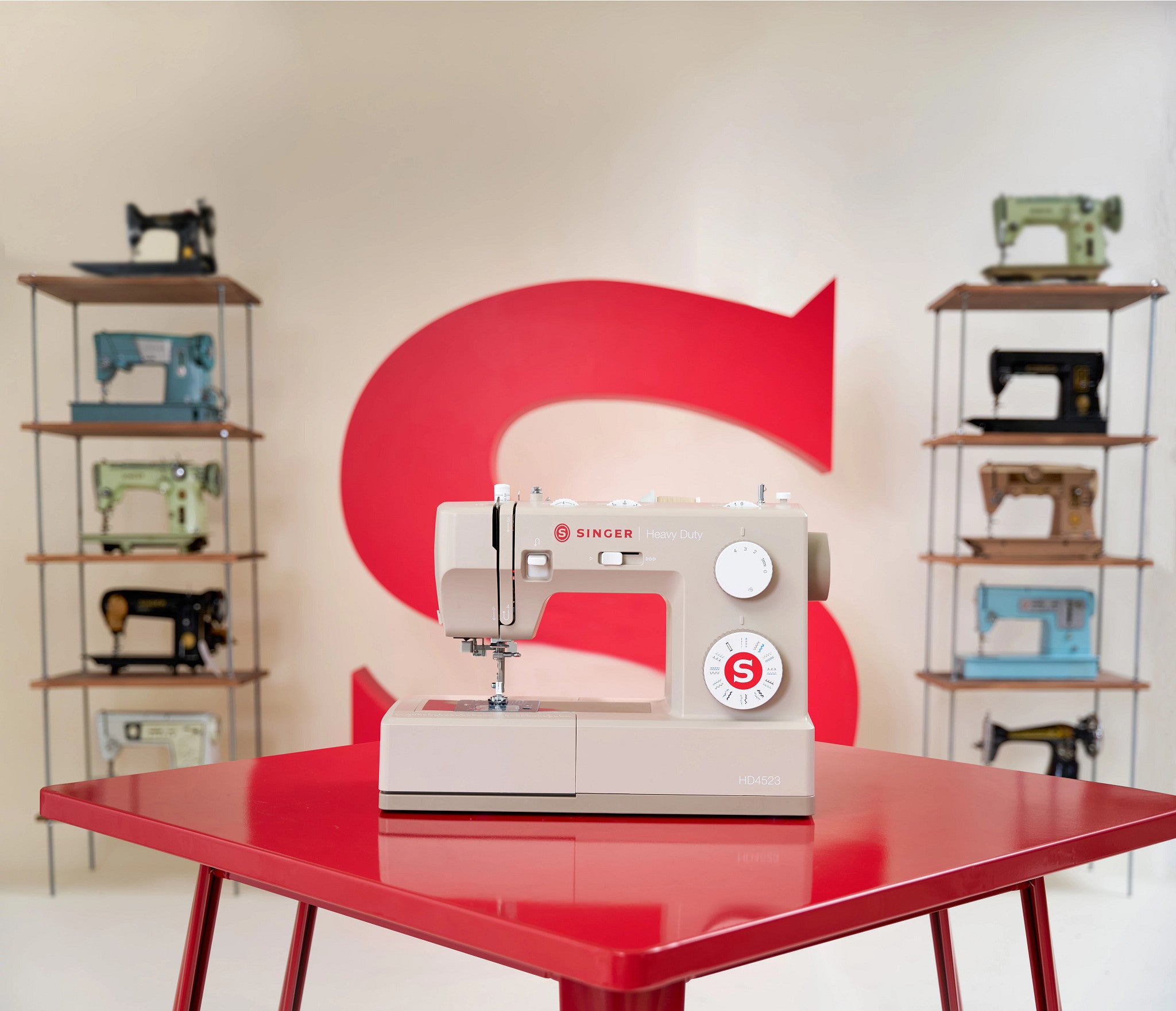 A beige and white Singer sewing machine sits on a glossy red table, with a large red stylized 'S' logo on the wall behind it and shelves of vintage sewing machines on both sides.