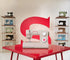A beige and white Singer sewing machine sits on a glossy red table, with a large red stylized 'S' logo on the wall behind it and shelves of vintage sewing machines on both sides.