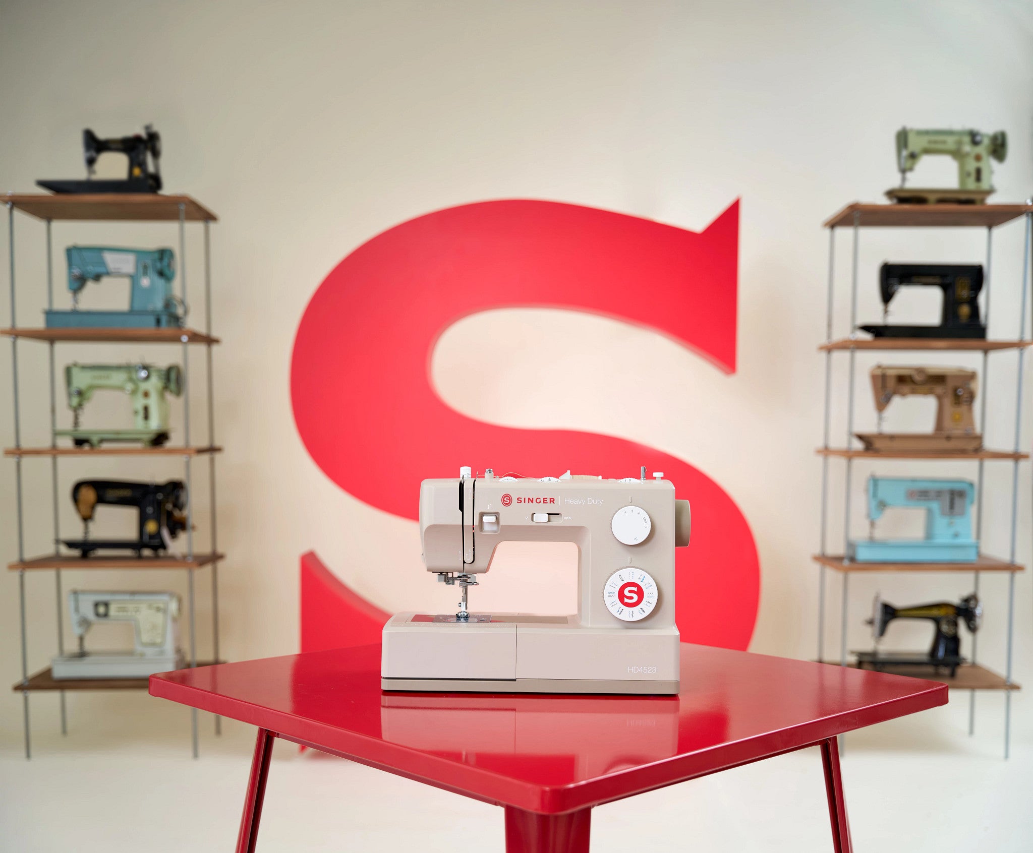 A light beige Singer sewing machine sits on a glossy red table, with a large red letter S on the wall behind it and shelves of other vintage sewing machines on both sides.