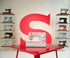 A light beige Singer sewing machine sits on a glossy red table, with a large red letter S on the wall behind it and shelves of other vintage sewing machines on both sides.